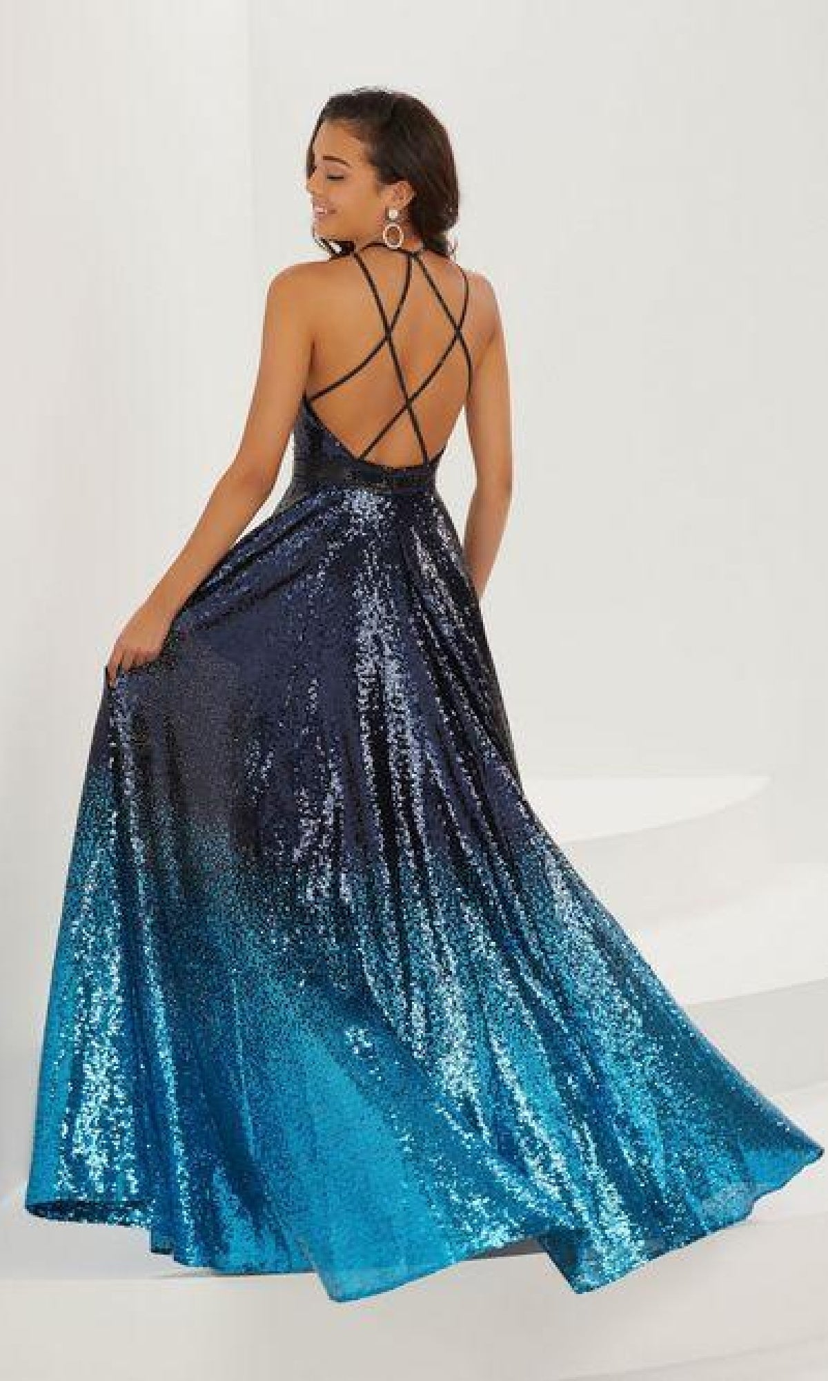 A woman poses in the A-Line Sequin Ombre Prom Dress: Tiffany 16941, featuring a sleeveless, backless design with a low V-neckline, thin crisscross straps, and a dark blue to light blue sequin gradient.