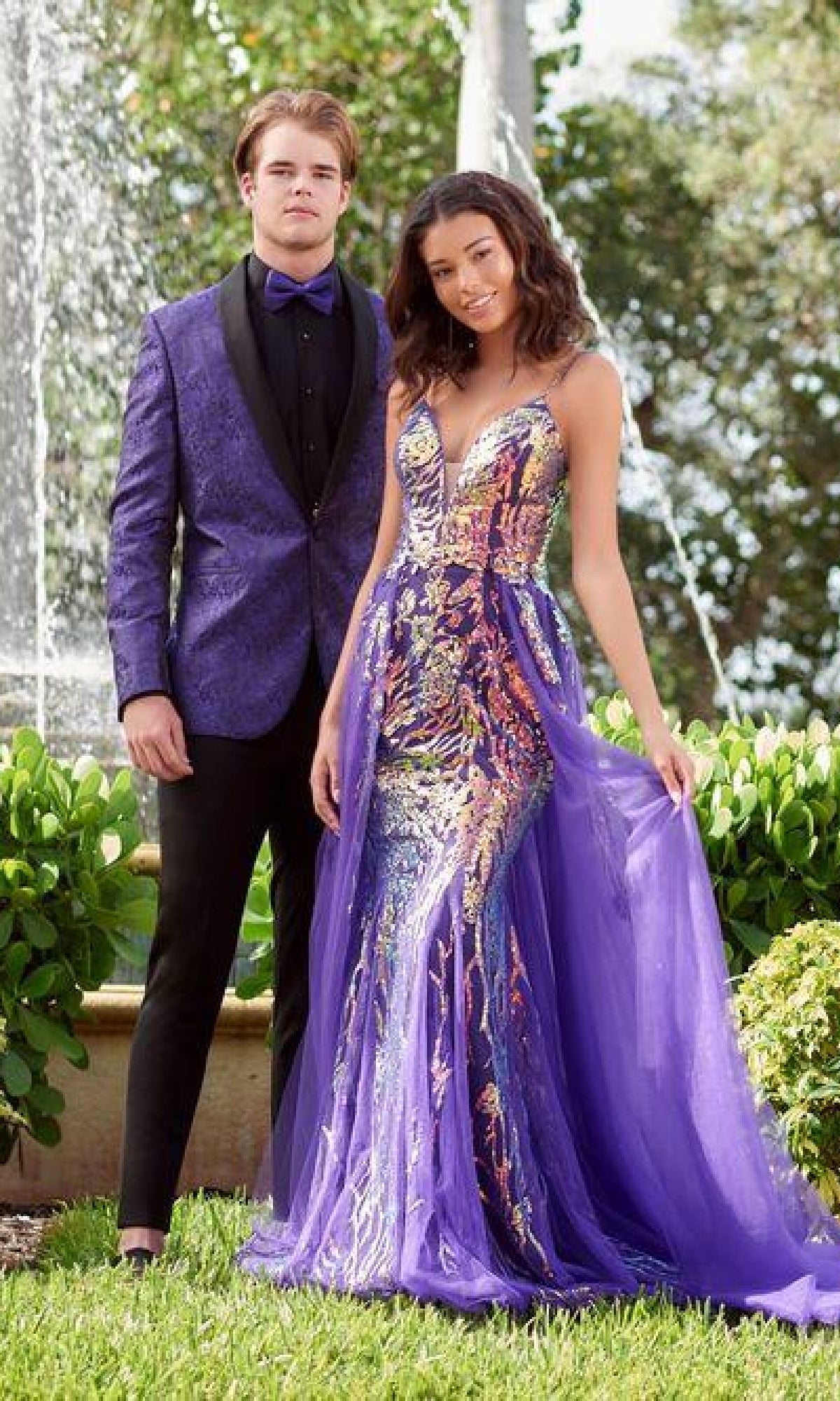 A young man in a purple tuxedo and a young woman wearing the Long Sequin Prom Dress: Tiffany 16944 with a tulle overlay pose outdoors by a fountain.