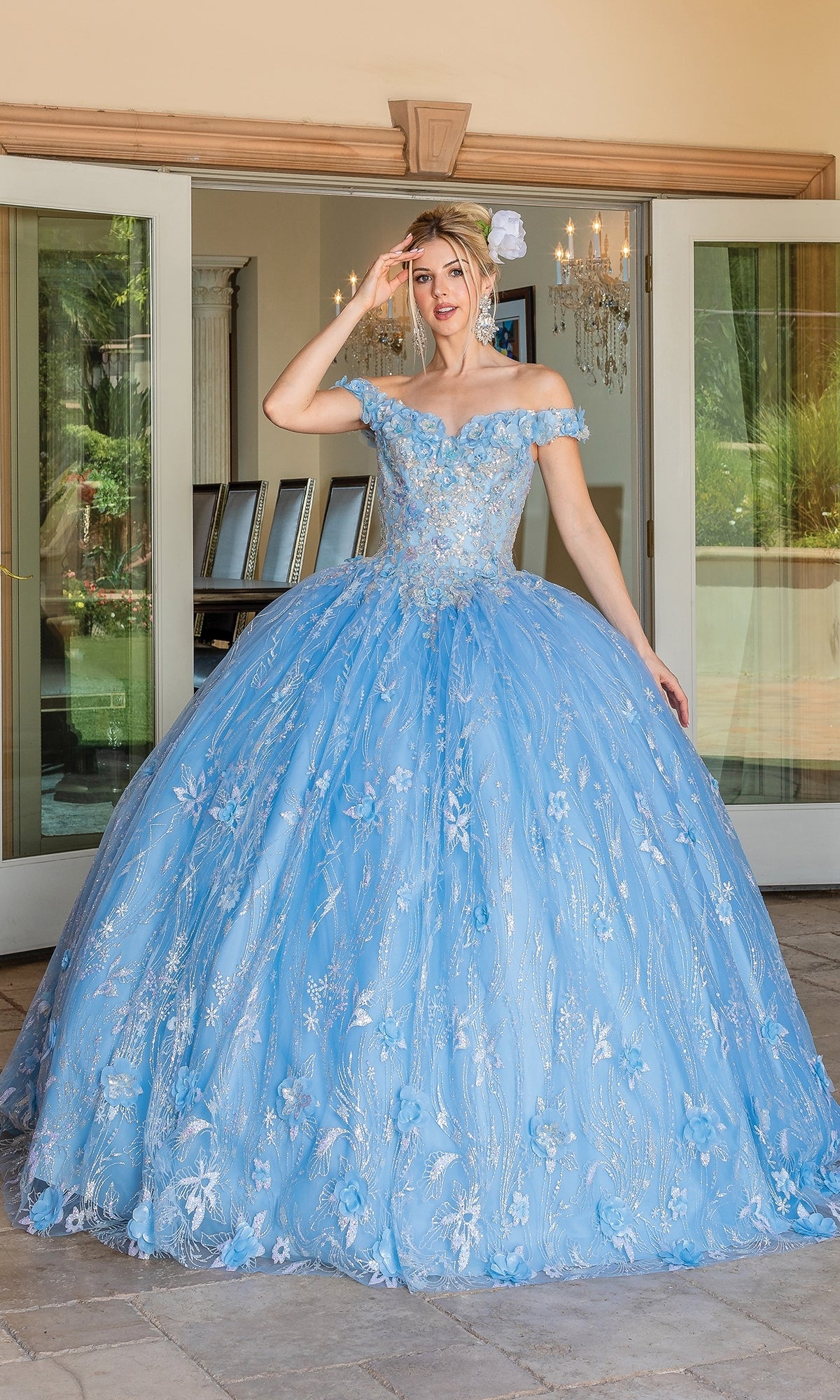 A woman stands outside in front of glass doors, wearing the Quinceanera Dress 1694 By Dancing Queen—an elegant off-the-shoulder ball gown with floral embroidery and a light blue full A-line skirt.