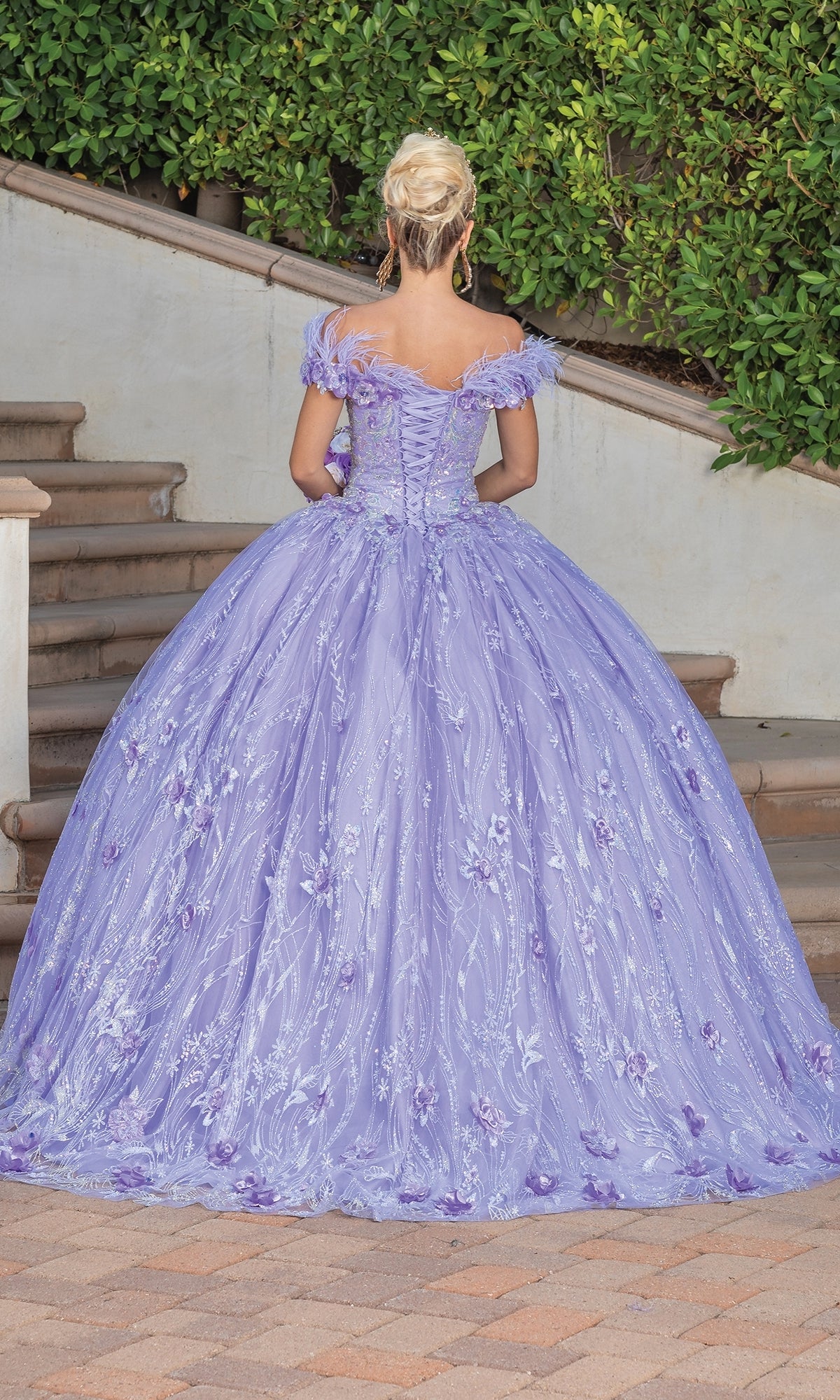 A person with blonde hair in an updo stands on outdoor steps wearing the Quinceanera Dress 1694 By Dancing Queen, a lilac off-the-shoulder ball gown with a full skirt and sparkling embellishments.