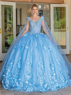 A woman in the Quinceanera Dress 1695 By Dancing Queen, featuring a beautiful Bahama Blue color and elegant cape sleeves, stands smiling in a doorway. Surrounded by chandeliers and lush greenery, delicate floral details accentuate her enchanting a-line ball gown look.