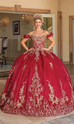 A woman stands indoors wearing Quinceanera Dress 1697 with Embroidery—an ornate, off-the-shoulder red gown featuring gold embroidery and a matching tiara.