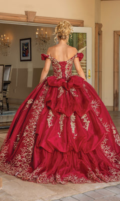 A person stands indoors wearing the Quinceanera Dress 1697 with Embroidery, a red off-the-shoulder gown featuring gold embroidery, layered ruffles, and a stunning sweetheart neckline, viewed from the back.