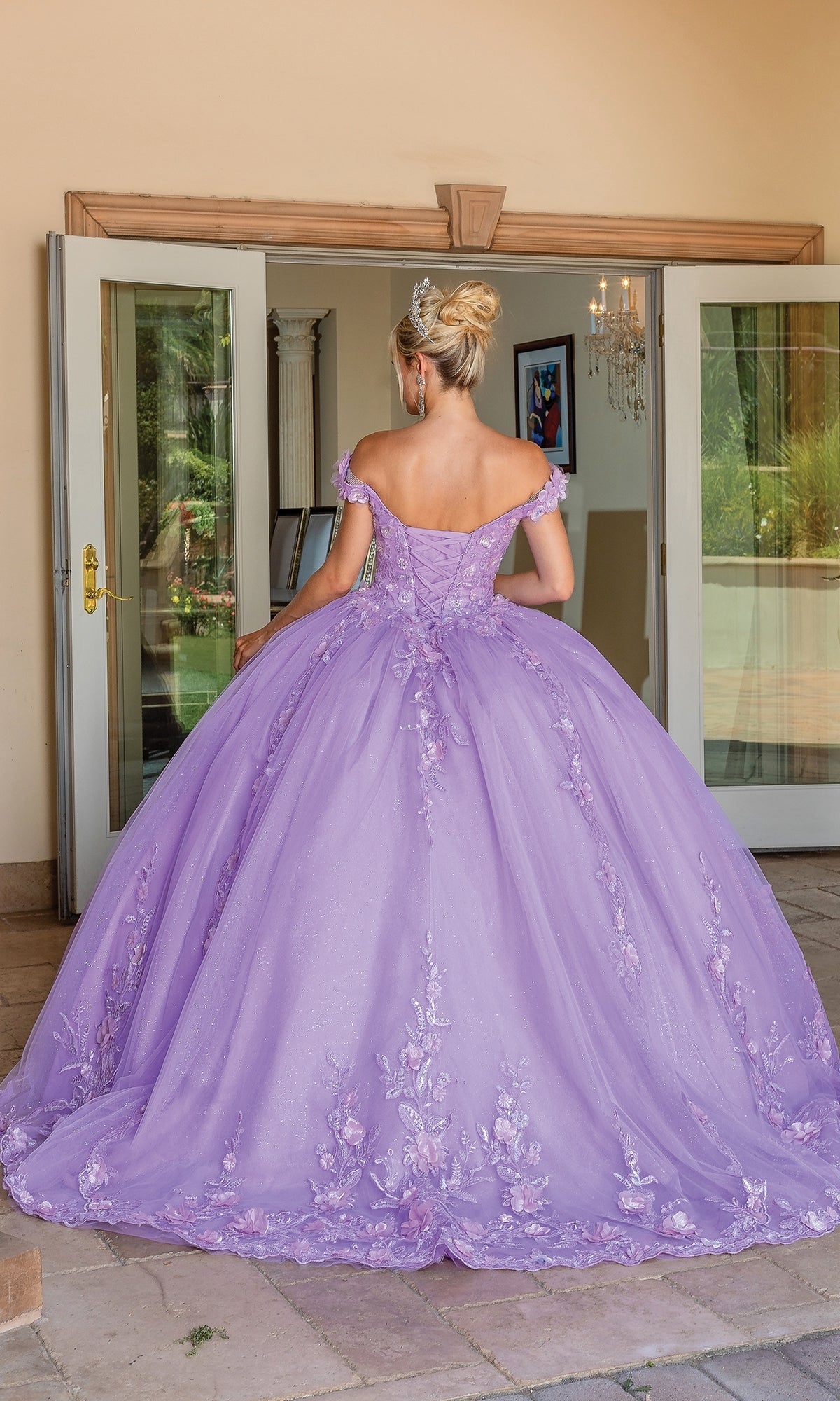 A woman in the Quinceanera Dress 1698 By Dancing Queen—a floor-length, off-the-shoulder gown with floral embroidery—stands before open double doors, her lavender ball gown elegantly trailing behind her.