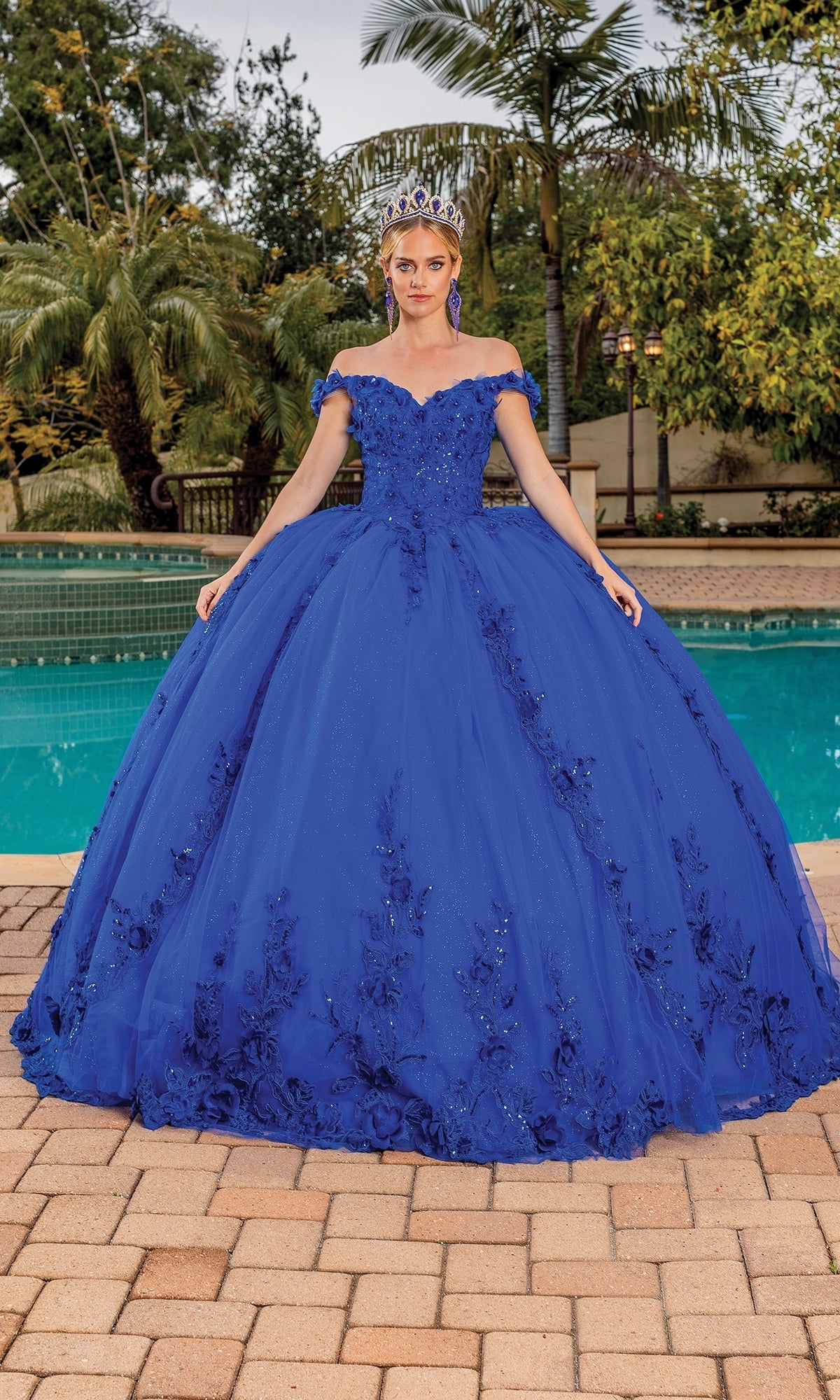 A woman stands by a pool on a tiled patio with palm trees, wearing the Quinceanera Dress 1698 By Dancing Queen—a stunning royal blue off-the-shoulder gown with floral details and a tiara.