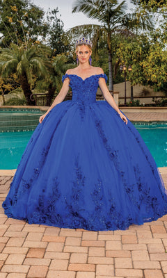 A woman stands by a pool on a tiled patio with palm trees, wearing the Quinceanera Dress 1698 By Dancing Queen—a stunning royal blue off-the-shoulder gown with floral details and a tiara.