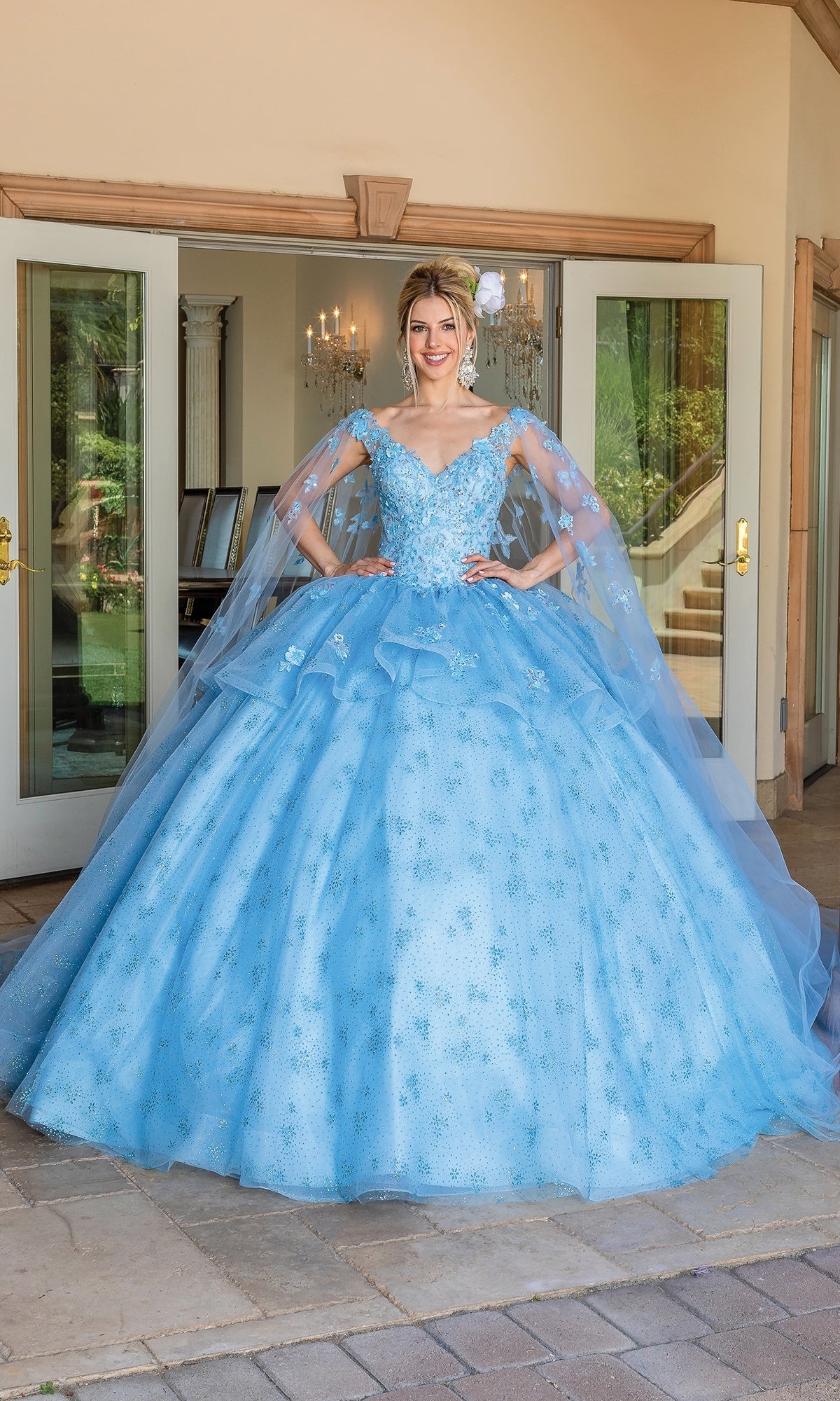 A woman stands outside double glass doors wearing the Quinceanera Dress 1702 By Dancing Queen, featuring a voluminous light blue ball gown with floral embroidery and a lace embellished cape.