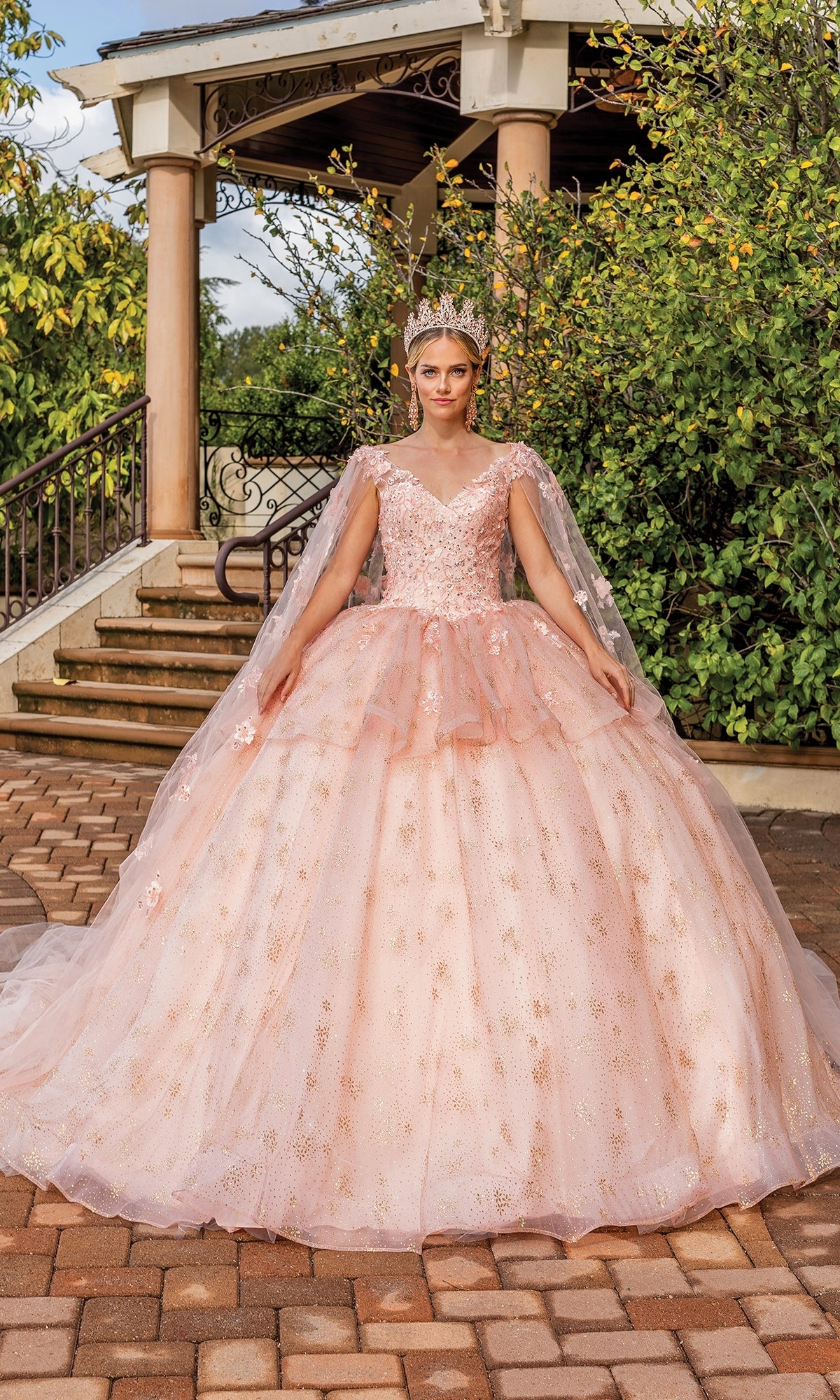 A young woman wearing the Quinceanera Dress 1702 By Dancing Queen and a tiara stands outdoors in front of a gazebo, posing on a paved walkway surrounded by greenery.