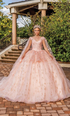 A young woman wearing the Quinceanera Dress 1702 By Dancing Queen and a tiara stands outdoors in front of a gazebo, posing on a paved walkway surrounded by greenery.