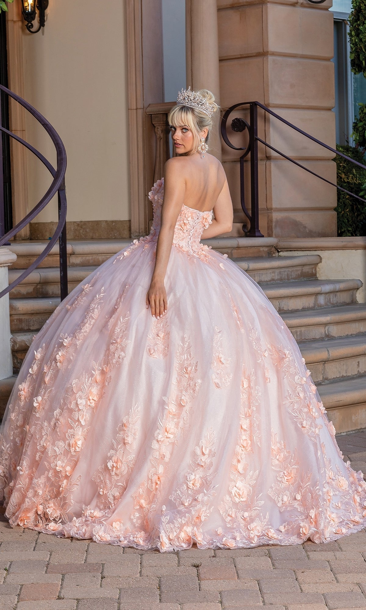 A woman wears the Pastel Peach Quinceanera Dress 1705 with a sweetheart strapless neckline and floral embroidery, standing on steps outside, silver tiara on her head as she looks over her shoulder.