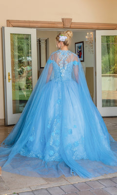 A woman wearing the Quinceanera Dress 1706 By Dancing Queen, a light blue ball gown with lace, tulle details, an off-the-shoulder design, and a flowing removable cape stands facing away in front of open double doors.