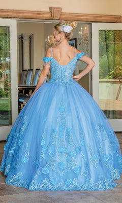 A woman faces away from the camera indoors, wearing the Quinceanera Dress 1706 by Dancing Queen—an ornate, light blue ball gown with floral embroidery and an off-the-shoulder design, standing in front of glass doors.