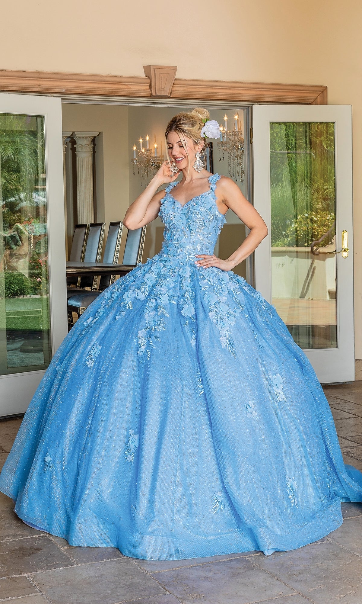A woman indoors smiles and holds her hair while wearing the Quinceanera Dress 1707 By Dancing Queen, a light blue ball gown with floral embroidery, with glass doors and a chandelier in the background.