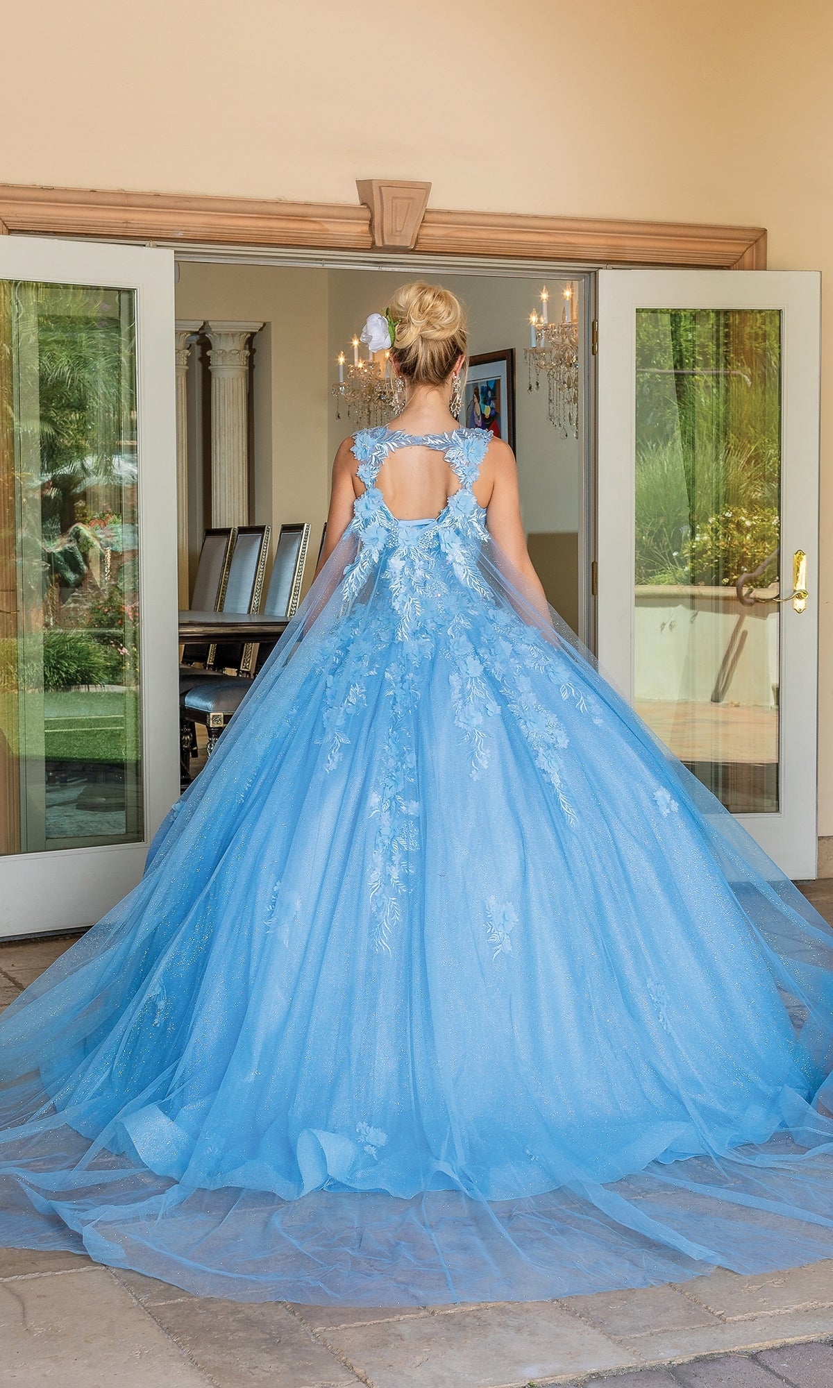 A person faces away from the camera in front of open glass doors, wearing the Quinceanera Dress 1707 By Dancing Queen—a floor-length blue ball gown with floral embroidery.