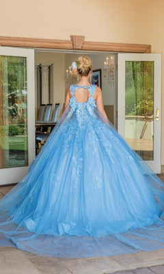 A person faces away from the camera in front of open glass doors, wearing the Quinceanera Dress 1707 By Dancing Queen—a floor-length blue ball gown with floral embroidery.