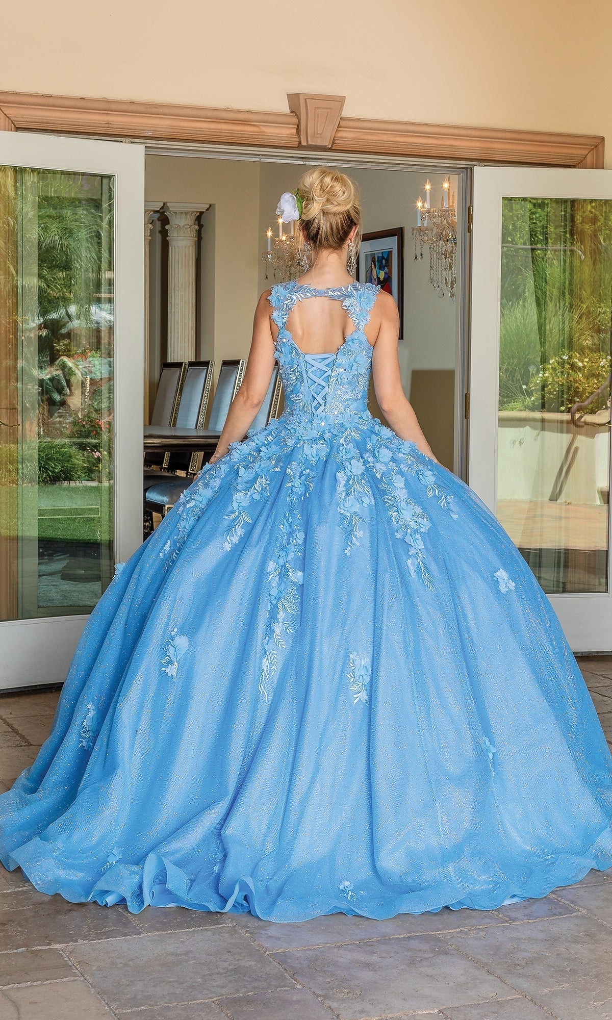 A woman outdoors faces glass doors in the Quinceanera Dress 1707 By Dancing Queen, a blue ball gown featuring a full skirt, lace-up back, and floral embroidered lace.