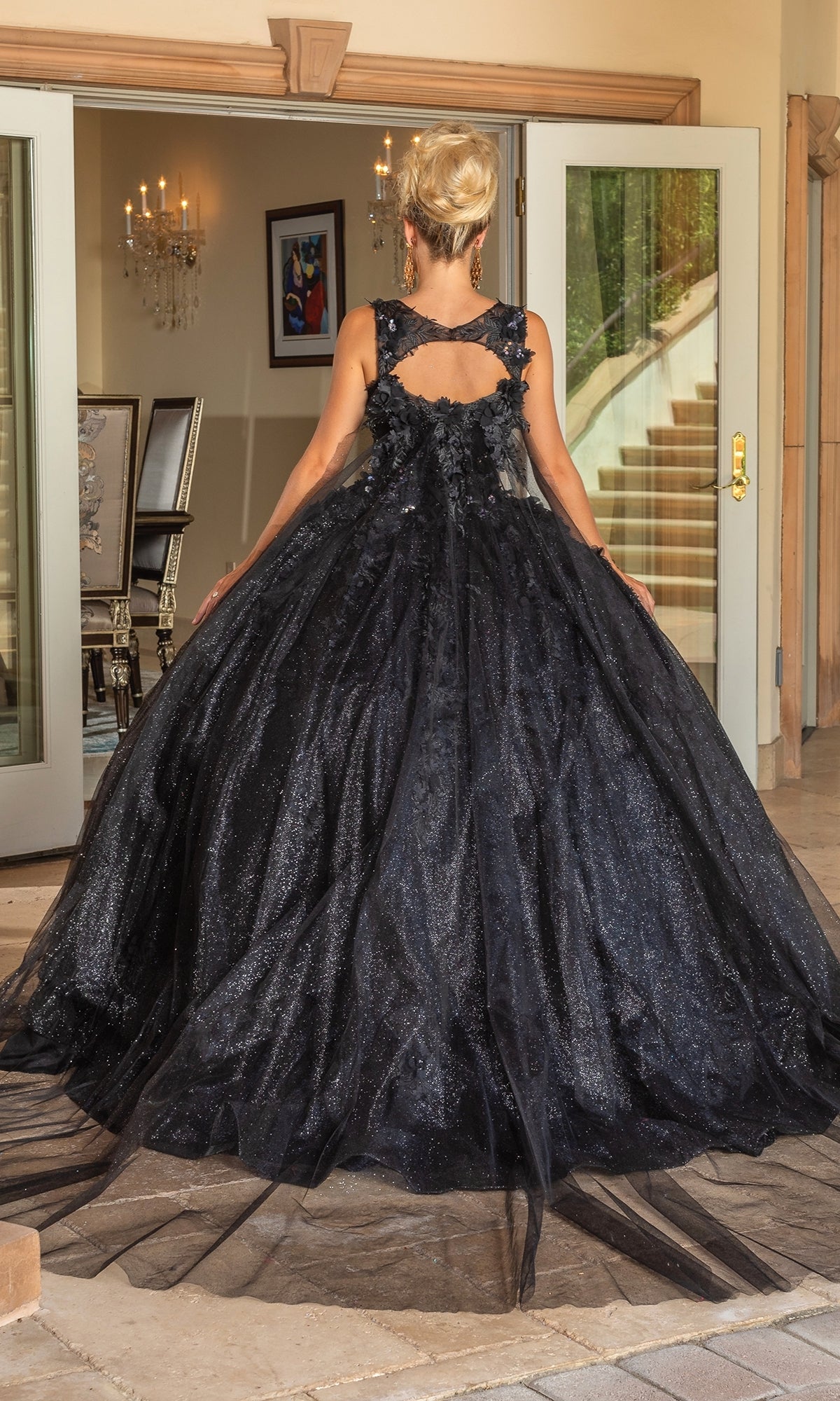 A person stands indoors facing away, wearing the floor-length black Quinceanera Dress 1707 By Dancing Queen featuring a cutout and floral embroidered back. Double doors and a staircase are visible in the background.