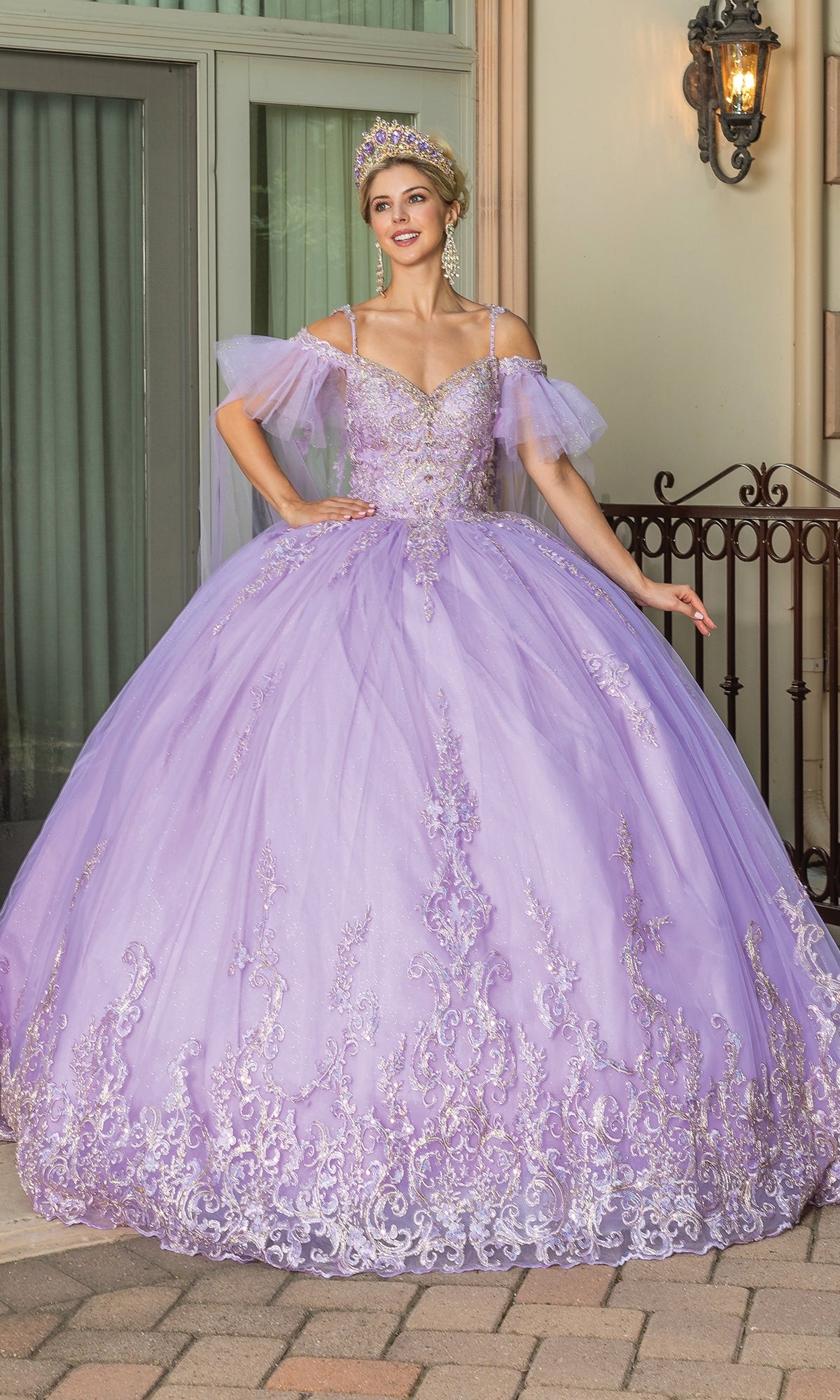 A woman poses gracefully outdoors in front of a wrought iron fence and window, wearing the Quinceanera Dress 1713 by Dancing Queen with floral embroidery, off-shoulder ruffle sleeves, and a jeweled tiara.