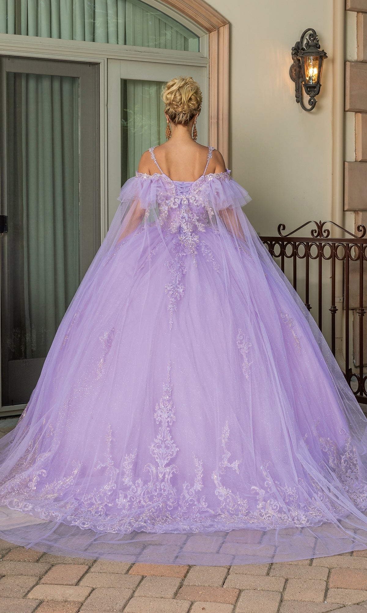 A woman, seen from behind, wears the Quinceanera Dress 1713 By Dancing Queen—a voluminous lavender gown with intricate embroidery and ruffle sleeves—outside a building with glass doors and a light fixture.