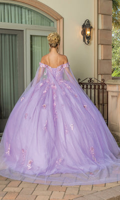 A woman with blonde hair in an updo stands outdoors with her back to the camera, wearing the Quinceanera Dress 1714 By Dancing Queen—an off-the-shoulder pastel purple ball gown adorned with floral appliqués.
