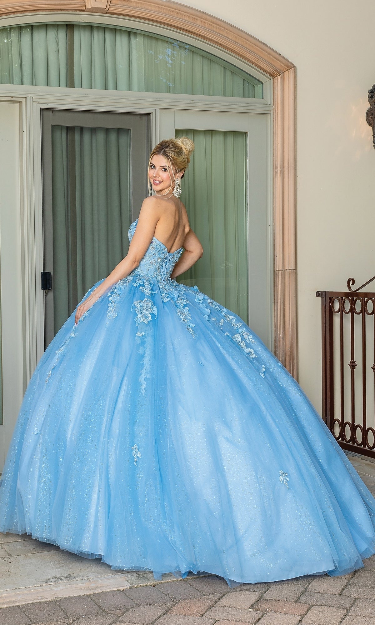 A woman wearing the Strapless Blue Quinceanera Dress 1717 with a sweetheart neckline stands outdoors in front of large glass doors, looking over her shoulder and smiling.