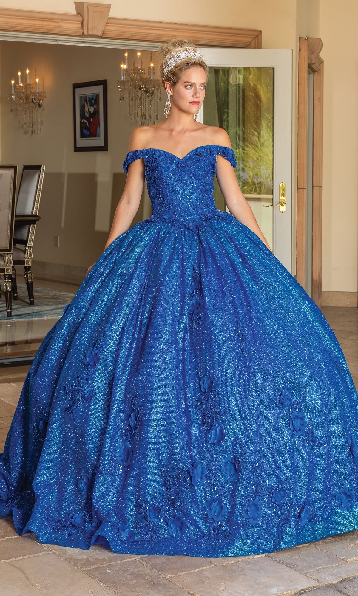 A woman stands indoors by open doors, wearing the sparkling Quinceanera Dress 1718 By Dancing Queen in royal blue with an off-the-shoulder, sweetheart neckline; a chandelier and dining table appear in the background.