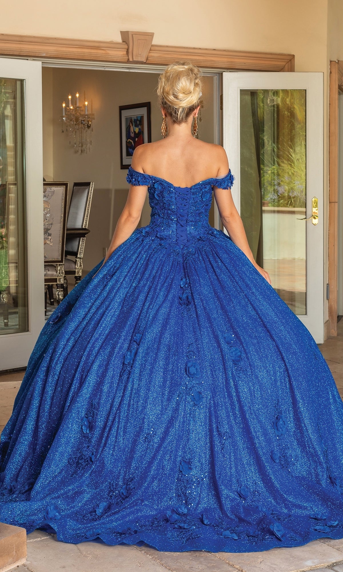 A woman stands indoors facing away, wearing the Quinceanera Dress 1718 By Dancing Queen—an off-the-shoulder, floor-length royal blue gown with a sparkly full skirt and lace-up back.