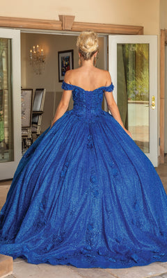 A woman stands indoors facing away, wearing the Quinceanera Dress 1718 By Dancing Queen—an off-the-shoulder, floor-length royal blue gown with a sparkly full skirt and lace-up back.