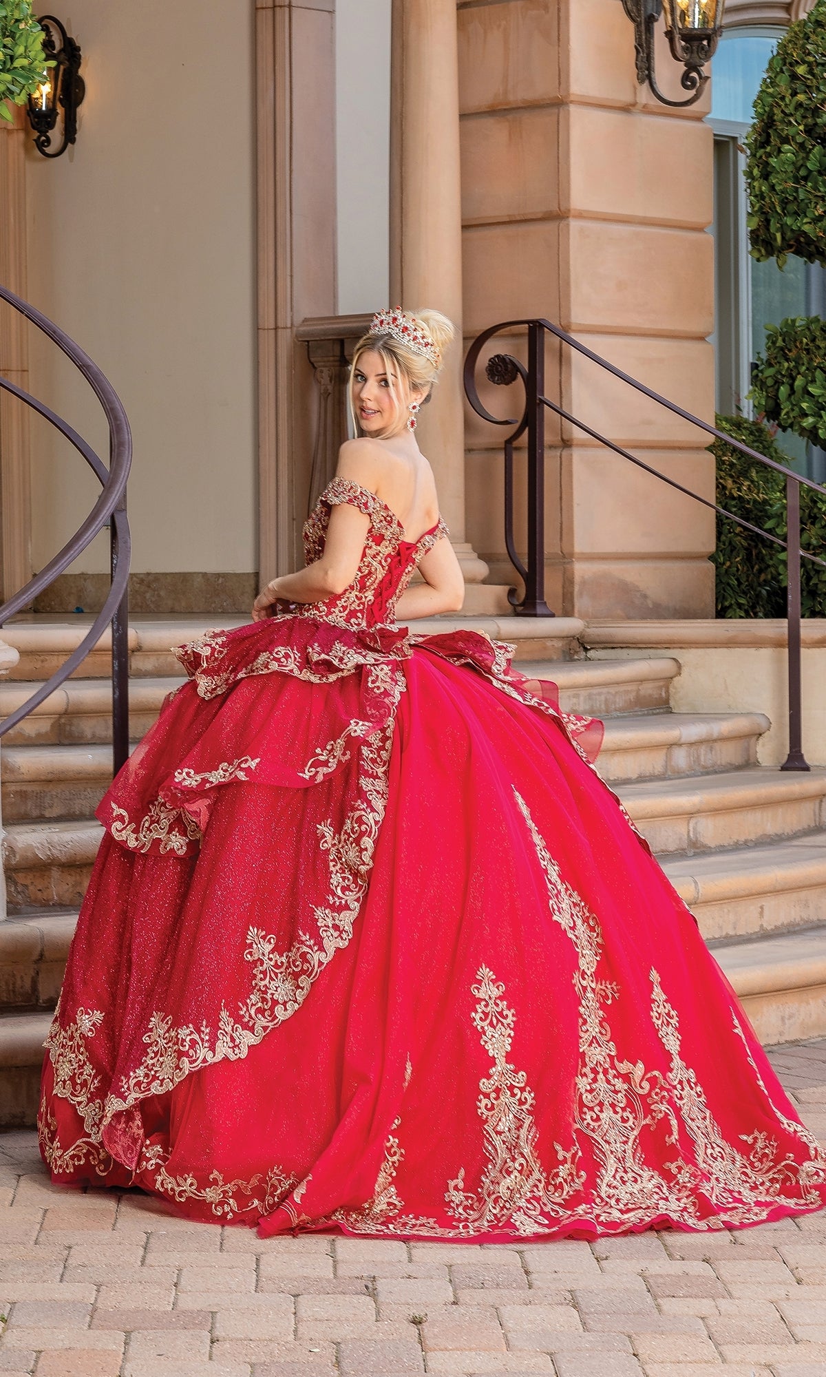 A young woman in the Quinceanera Dress 1720 by Dancing Queen, a bright red off-the-shoulder gown with gold embroidery, stands on a stone patio, glancing over her shoulder at the camera.