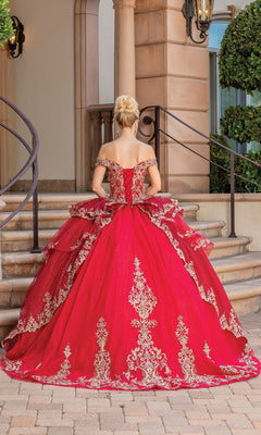 A woman wearing the Quinceanera Dress 1720 By Dancing Queen, an off-the-shoulder gold ball gown with lace embroidery, stands on a cobblestone path before stone steps and ornate railings, facing away from the camera.