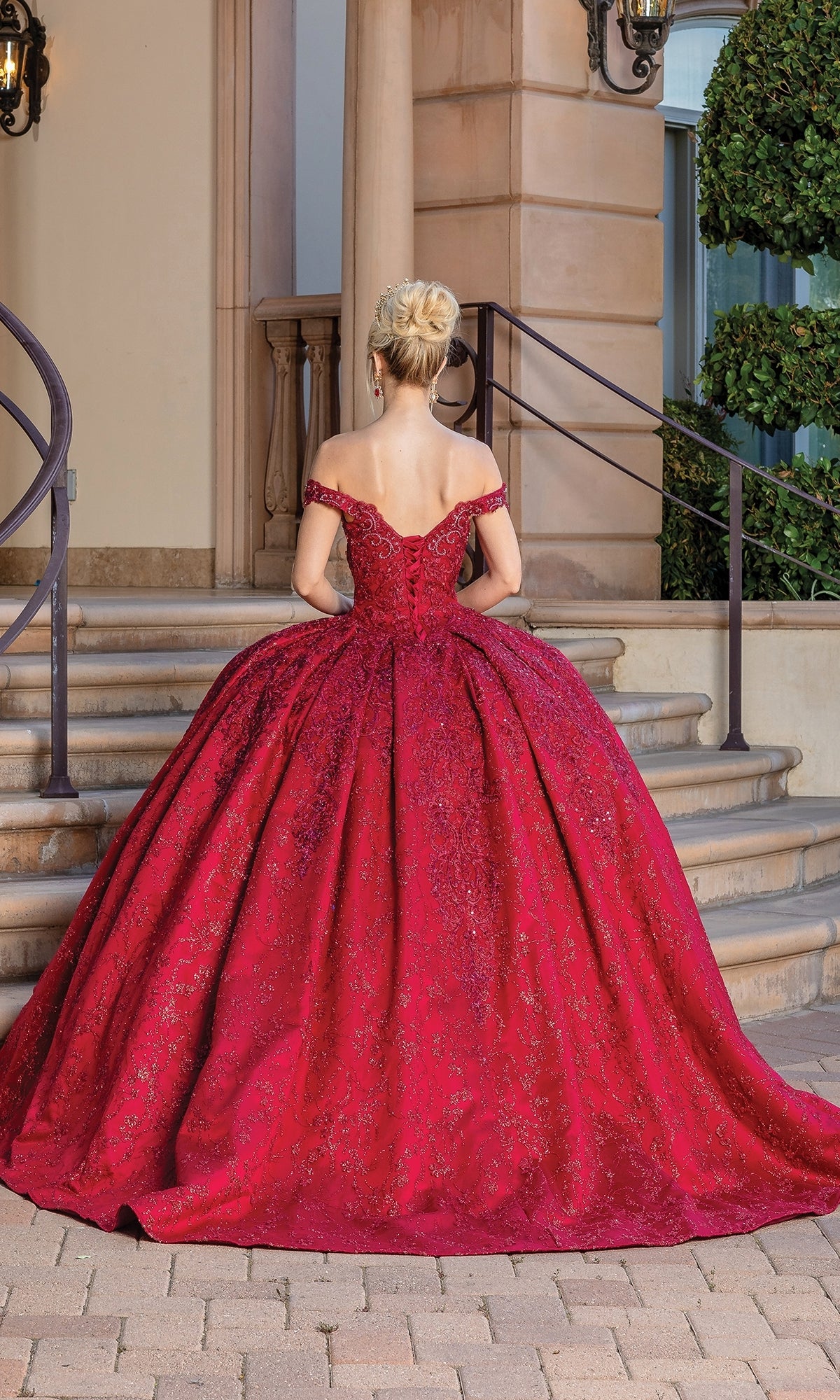 A woman stands with her back to the camera in front of stone steps and a building, wearing the Burgundy Red Quinceanera Dress 1722—an off-the-shoulder ball gown featuring a full skirt.
