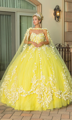A woman stands outdoors near a window beneath a wall lantern, wearing the Quinceanera Dress 1725 by Dancing Queen—a yellow A-line gown with a sweetheart strapless neckline and removable cape.