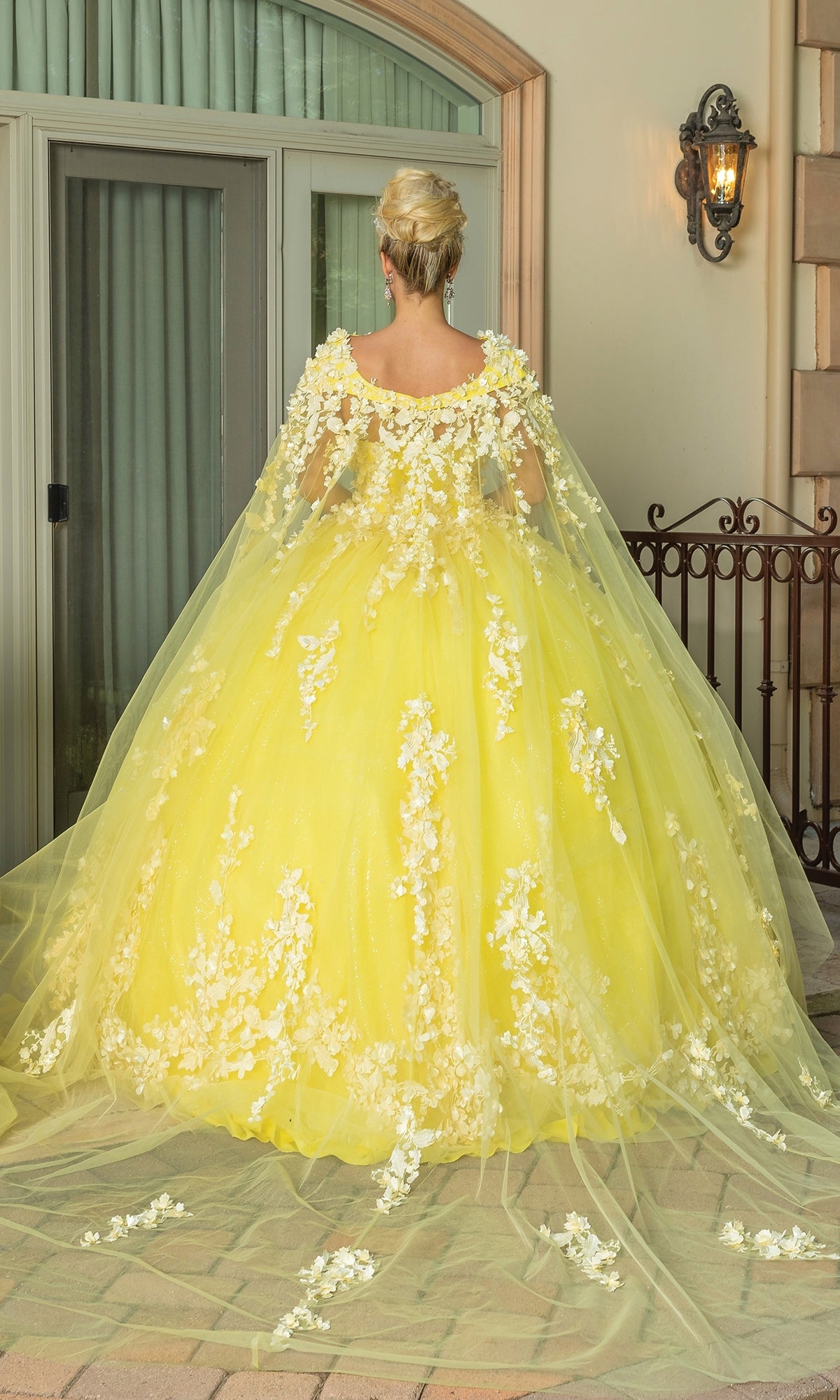 A woman faces away from the camera, wearing the Quinceanera Dress 1725 By Dancing Queen—a yellow a-line gown with white floral appliqués, a strapless sweetheart neckline, and removable sheer cape—outside a building with glass doors and lantern.