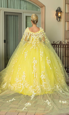 A woman faces away from the camera, wearing the Quinceanera Dress 1725 By Dancing Queen—a yellow a-line gown with white floral appliqués, a strapless sweetheart neckline, and removable sheer cape—outside a building with glass doors and lantern.
