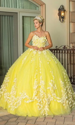 A woman stands outdoors wearing the Quinceanera Dress 1725 By Dancing Queen—a yellow a-line gown with floral appliqués, a strapless sweetheart neckline, voluminous skirt, headband accessory, and a removable cape for added flair.