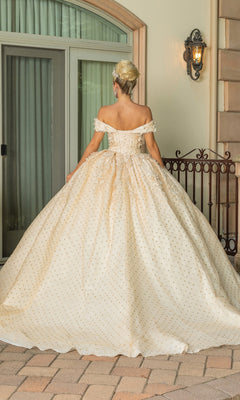 A woman stands facing away before a glass door and iron fence, wearing the Quinceanera Dress 1726 By Dancing Queen—an off-the-shoulder gown with a full ivory skirt accented by gold dots.