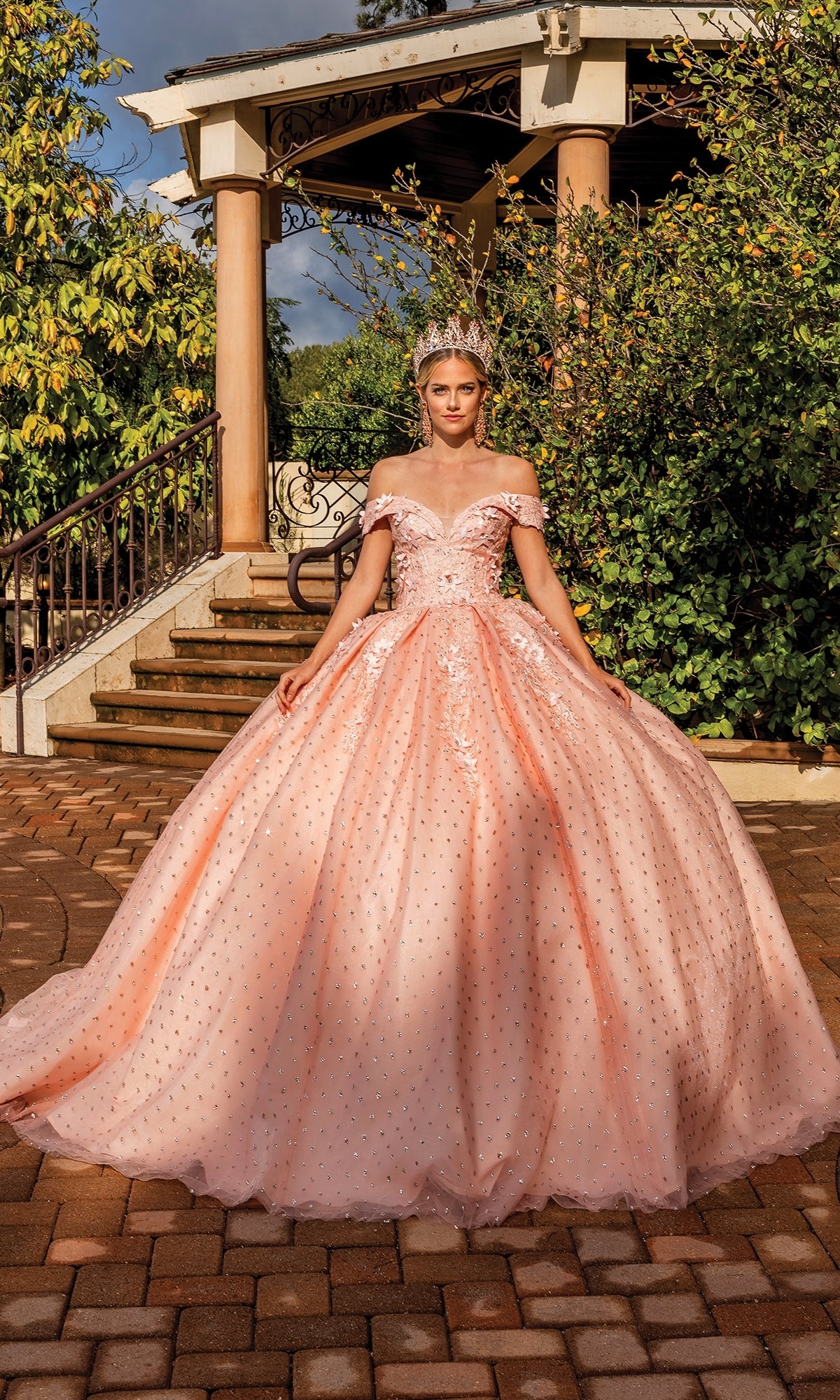 A young woman wearing the Quinceanera Dress 1726 By Dancing Queen with a tiara stands outdoors in front of a gazebo and greenery.