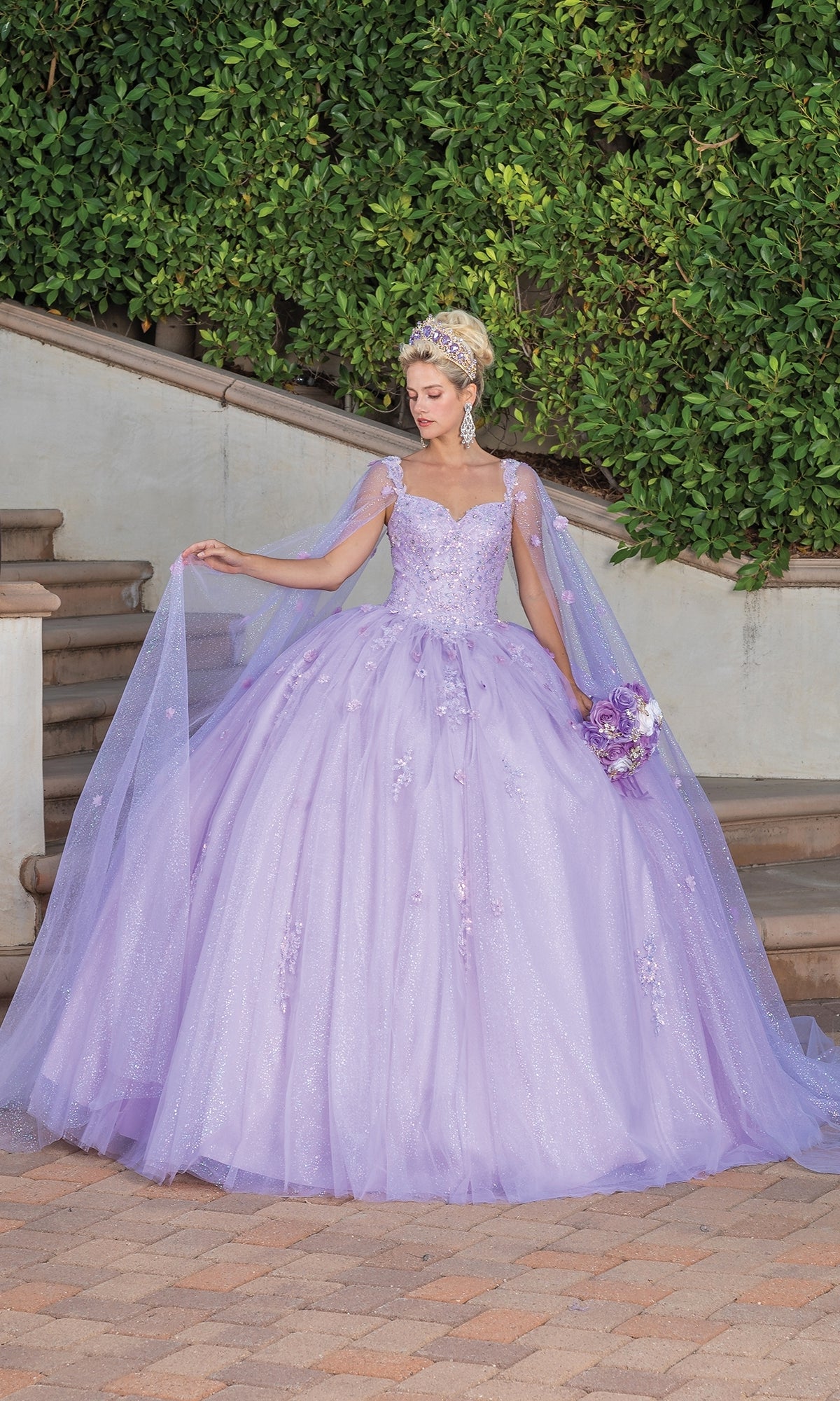 A woman stands outdoors in the Quinceanera Dress 1731 By Dancing Queen, a lilac ball gown with sparkly tulle and a sweetheart neckline, holding a matching bouquet amid greenery and stone steps.