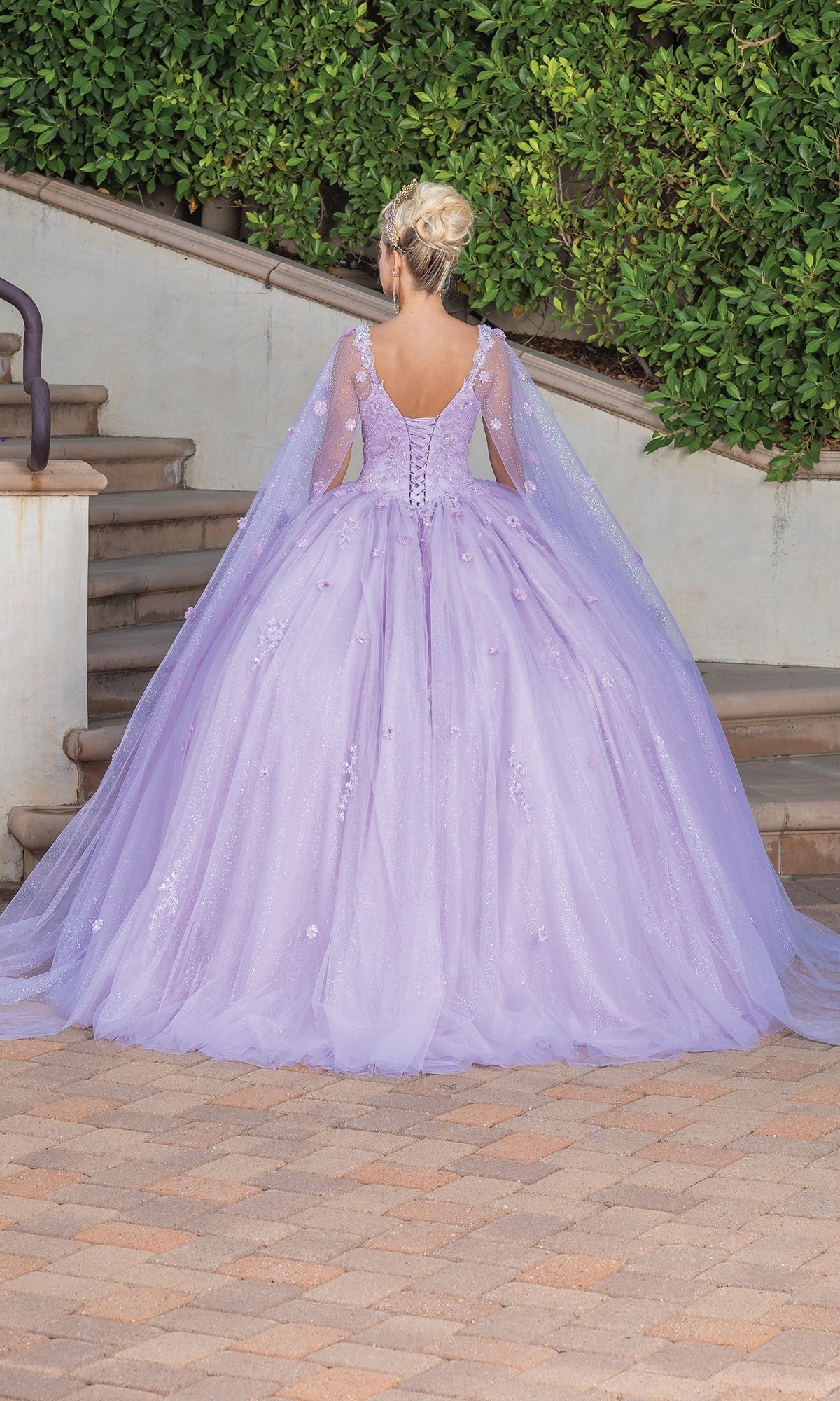 A person with blonde hair in an updo wears the Quinceanera Dress 1731 By Dancing Queen—a lilac ball gown with a full skirt, sheer cape, and sweetheart neckline—while standing on a brick path near stairs and greenery.