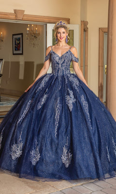 A woman stands indoors wearing the Quinceanera Dress 1732 By Dancing Queen—an off-the-shoulder, dark blue A-line ball gown adorned with silver lace embroidery and a tiara.