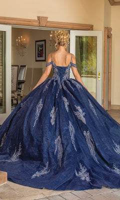 A woman wearing the Quinceanera Dress 1732 By Dancing Queen, an off-the-shoulder navy blue A-line ball gown with silver lace embroidery, stands with her back to the camera holding out the skirt in a room with open glass doors.