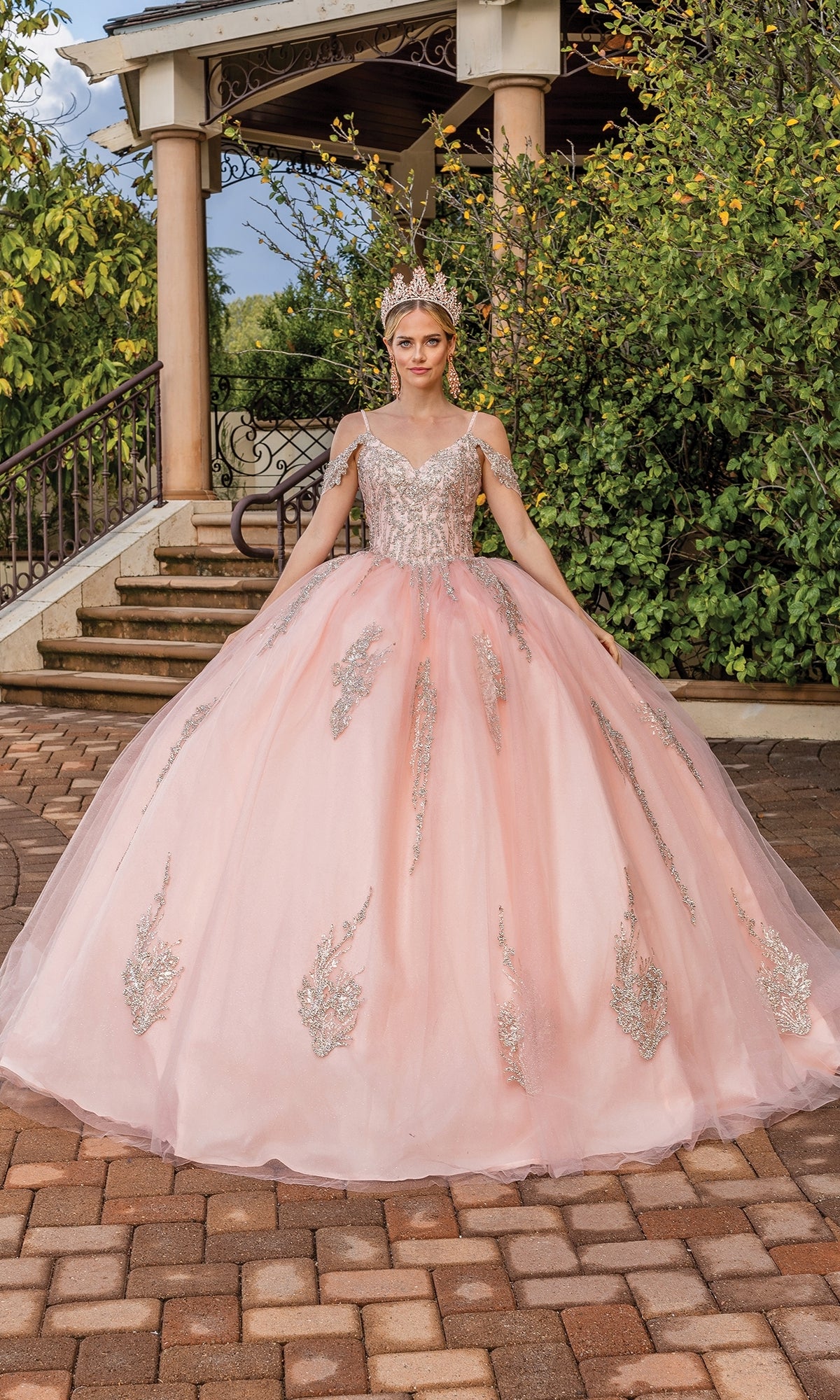 A woman stands outdoors on a brick pathway in front of steps and greenery, wearing the Quinceanera Dress 1732 By Dancing Queen—a pink off-the-shoulder A-line ball gown with silver embellishments, lace embroidery, and a tiara.