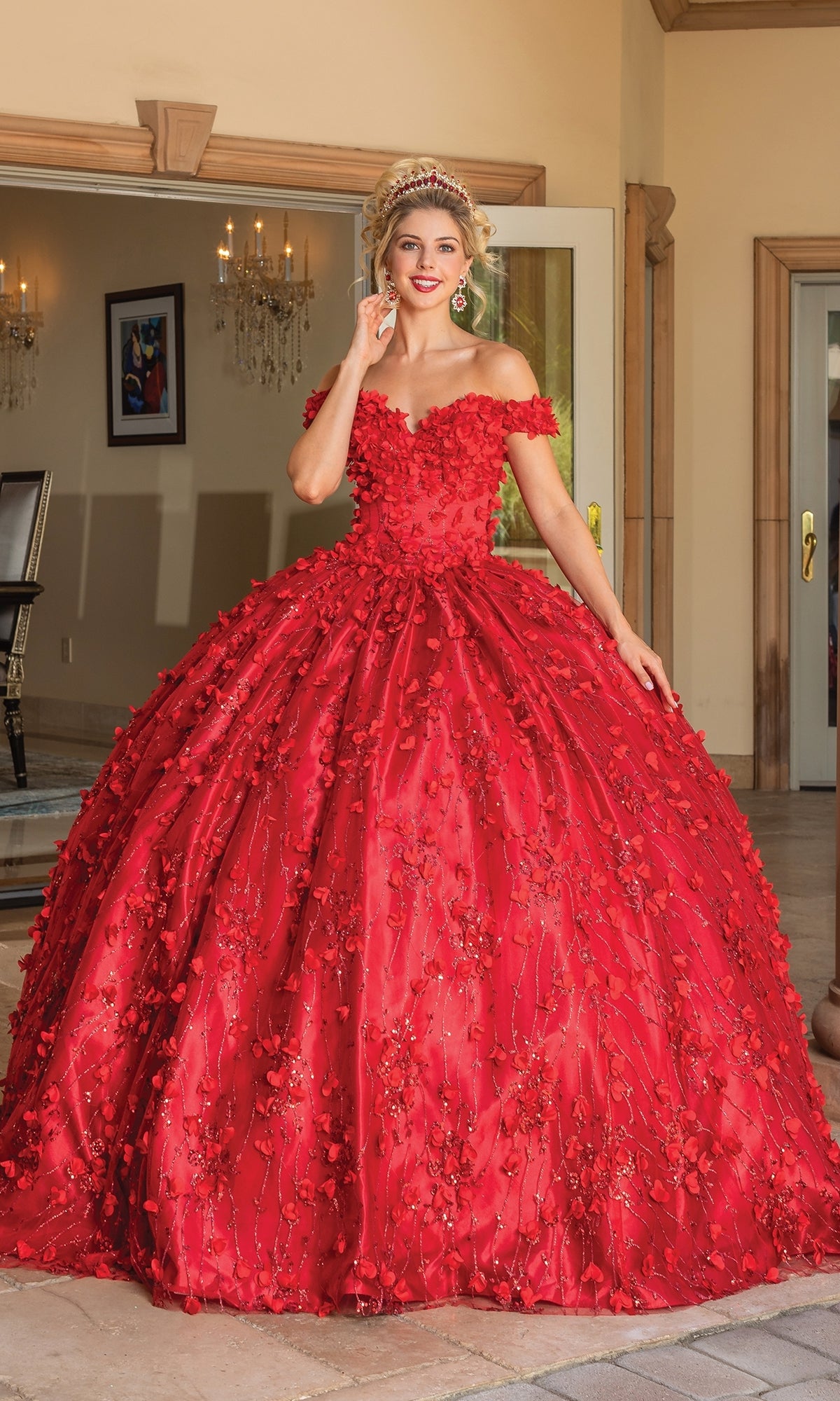 A person wearing the Quinceanera Dress 1734 By Dancing Queen, an off-the-shoulder, a-line ball gown with floral embellishments, smiles at the camera while standing indoors near open glass doors.