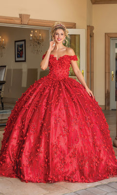 A person wearing the Quinceanera Dress 1734 By Dancing Queen, an off-the-shoulder, a-line ball gown with floral embellishments, smiles at the camera while standing indoors near open glass doors.