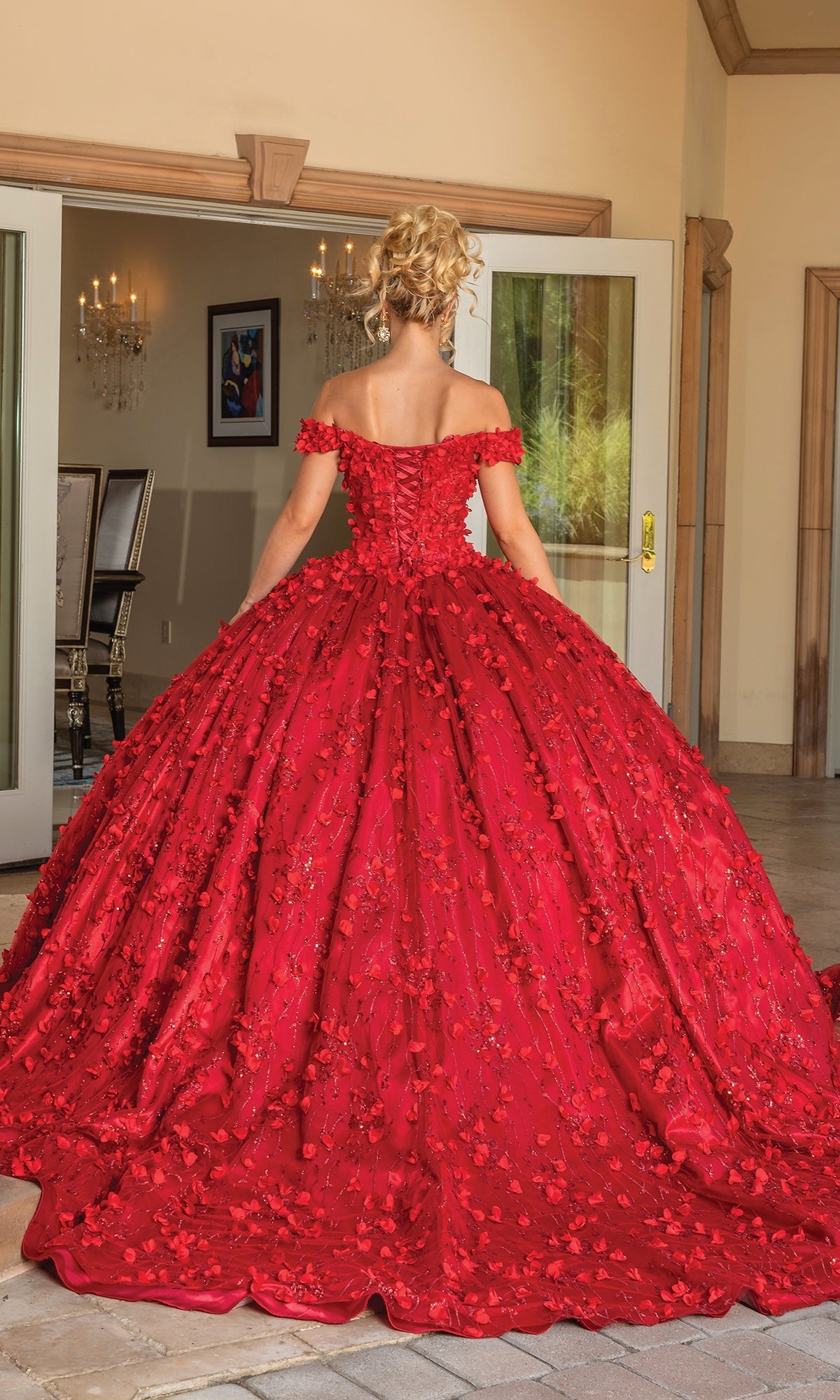 A woman with blonde hair in an updo wears the Quinceanera Dress 1734 By Dancing Queen, a red off-shoulder a-line ball gown with floral details, as she stands indoors before open glass doors.
