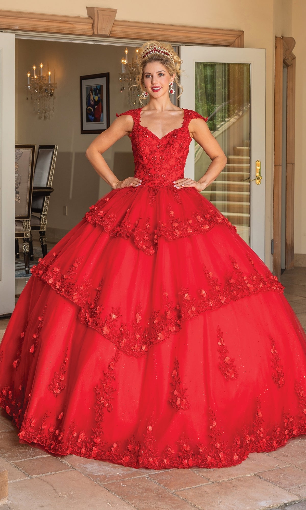 A woman stands indoors, hands on hips, wearing the Quinceanera Dress 1735 By Dancing Queen—a striking red A-line ball gown featuring floral appliqués, cap sleeves, and a layered tulle skirt.