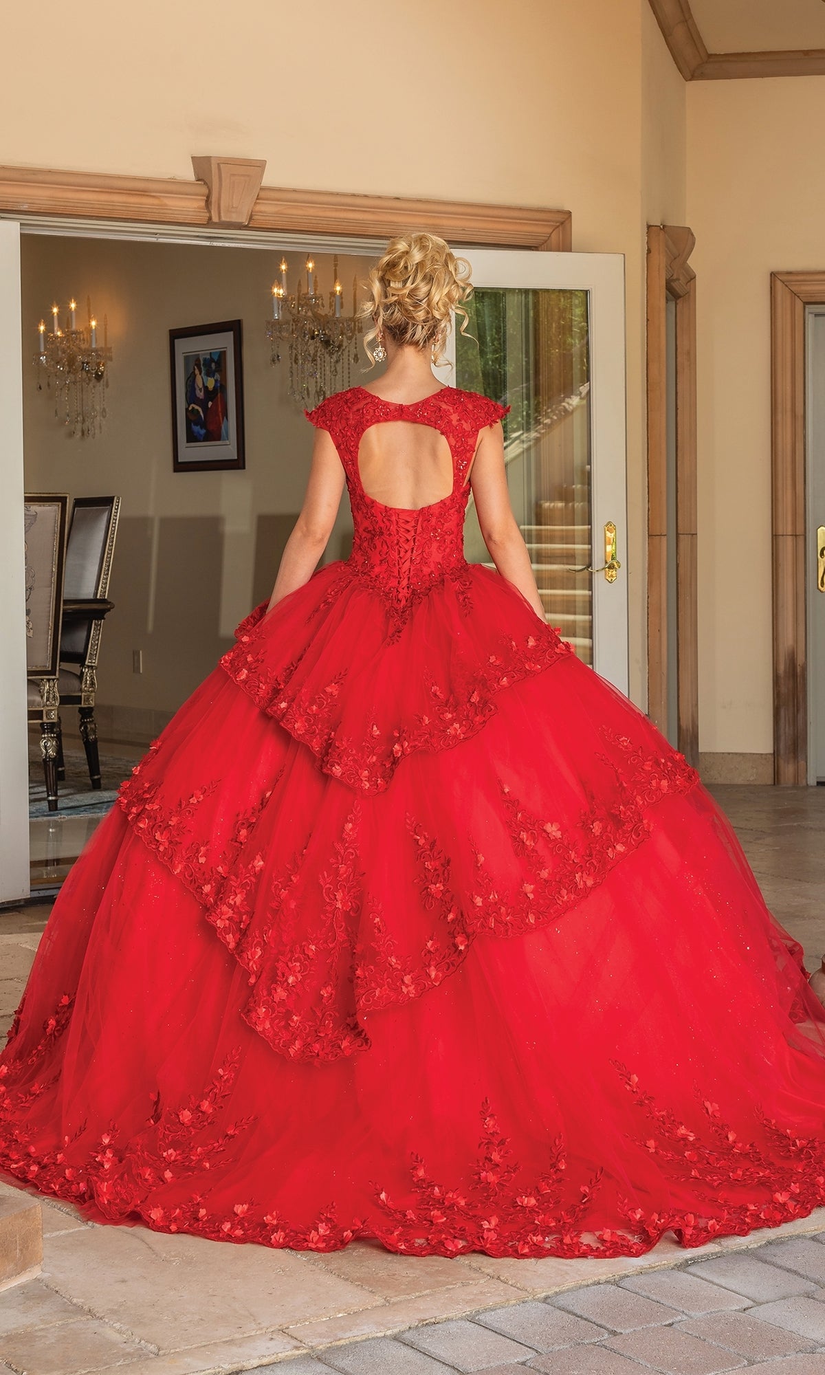 A woman stands indoors, facing away in the Quinceanera Dress 1735 By Dancing Queen—a voluminous red A-line ball gown with lace details and a keyhole back.
