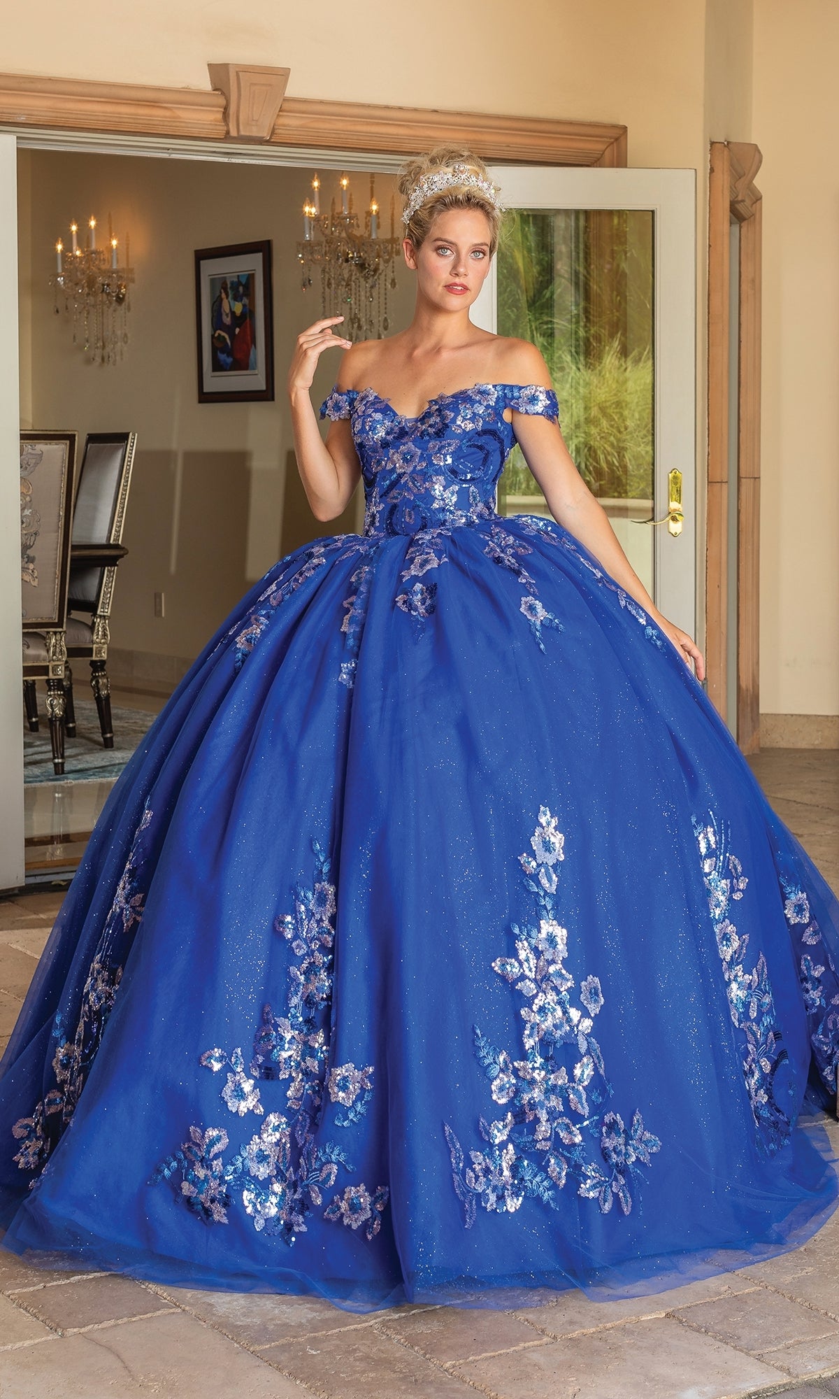 A woman stands indoors wearing the Quinceanera Dress 1737 By Dancing Queen, a royal blue ball gown with silver floral patterns, a full skirt, and an elegant off the shoulder design.