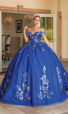 A woman stands indoors wearing the Quinceanera Dress 1737 By Dancing Queen, a royal blue ball gown with silver floral patterns, a full skirt, and an elegant off the shoulder design.