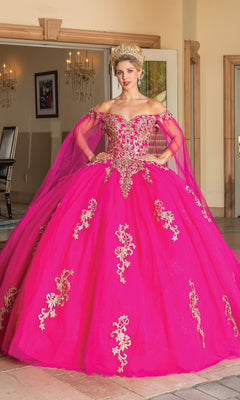 A woman wears the Quinceanera Dress 1740 by Dancing Queen, a bright pink off-shoulder gown with gold embroidery and tulle, indoors. She accessorizes with a jeweled tiara and looks toward the camera.